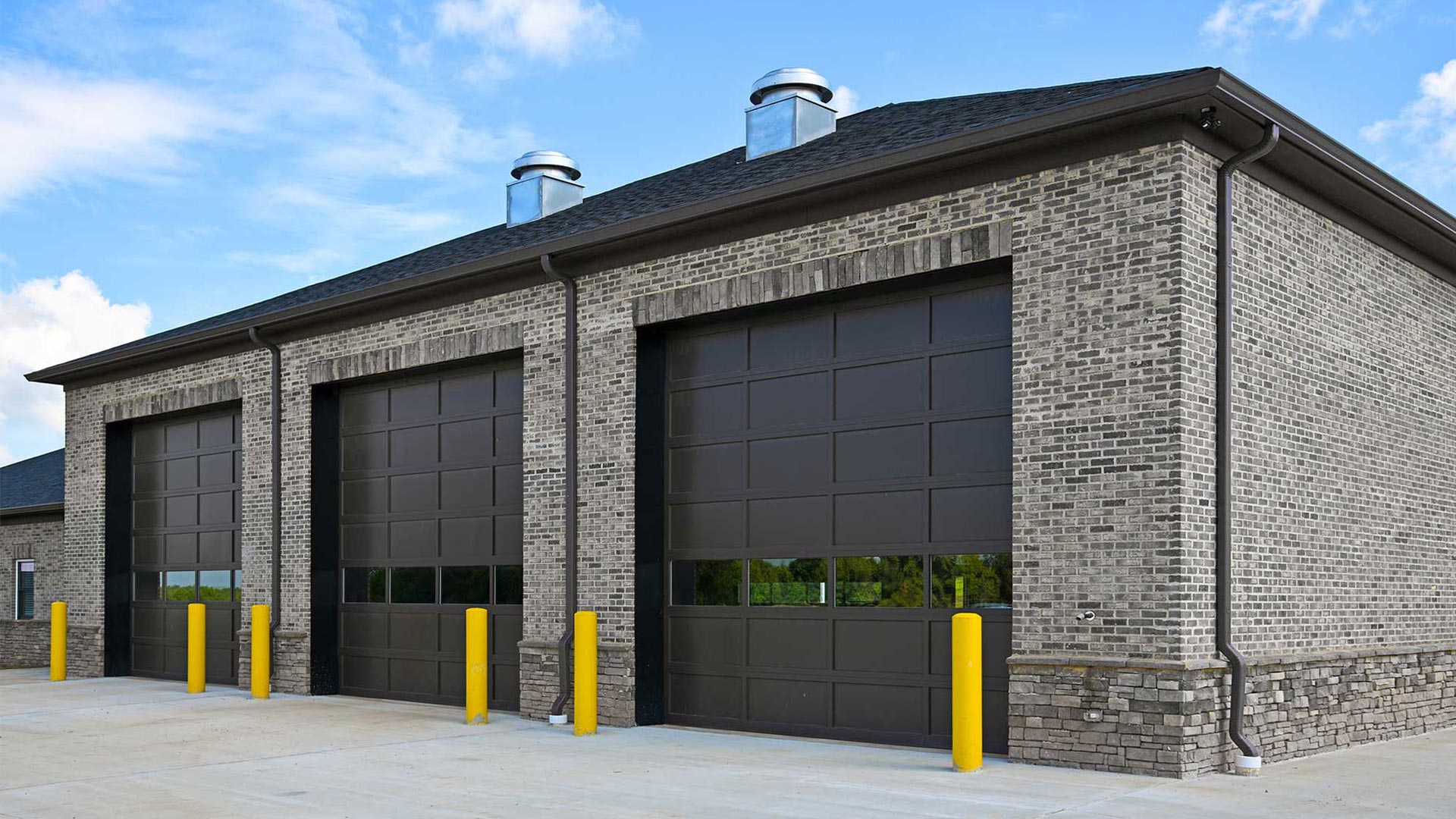 Three dark gray overhead doors with rows of window panels on a commercial garage with stone veneer