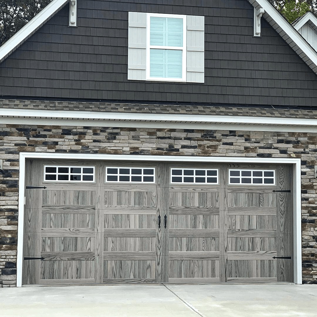 Gray wood garage doors with four window panes, each with a grille, on suburban home with stone veneer and dark gray siding